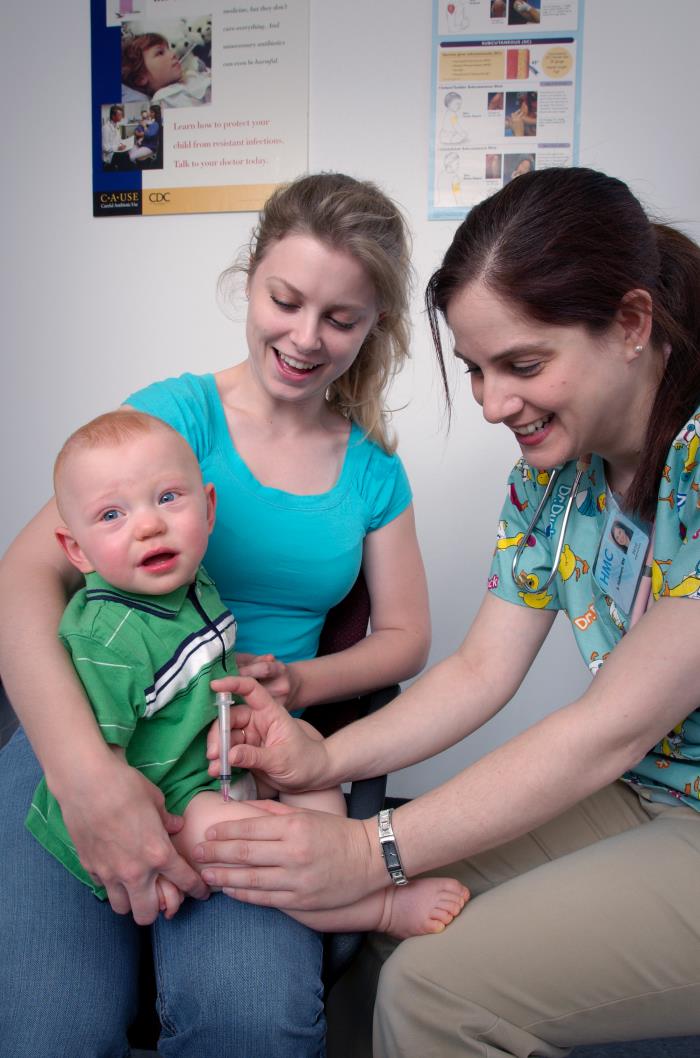 An infant is held on his mother's lap while getting an intramuscular injection in his thigh by a nurse.